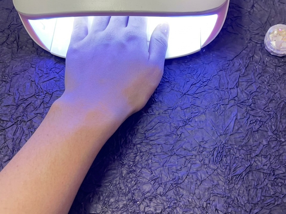 Una mano descansando dentro de una brillante lámpara de uñas UV/LED para curar esmalte de gel, sentada sobre una superficie morada texturizada.