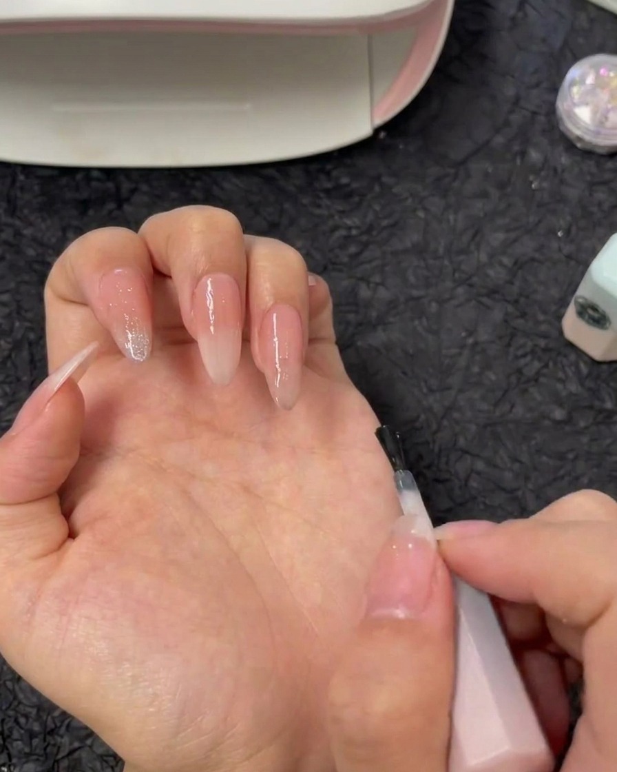Un pincel aplicando gel de refuerzo transparente directamente sobre una uña índice con base degradada nude.