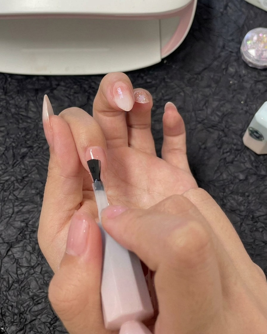 Una mano aplicando una capa de gel constructor transparente sobre la uña del dedo anular usando un pincel de esmalte de gel.
