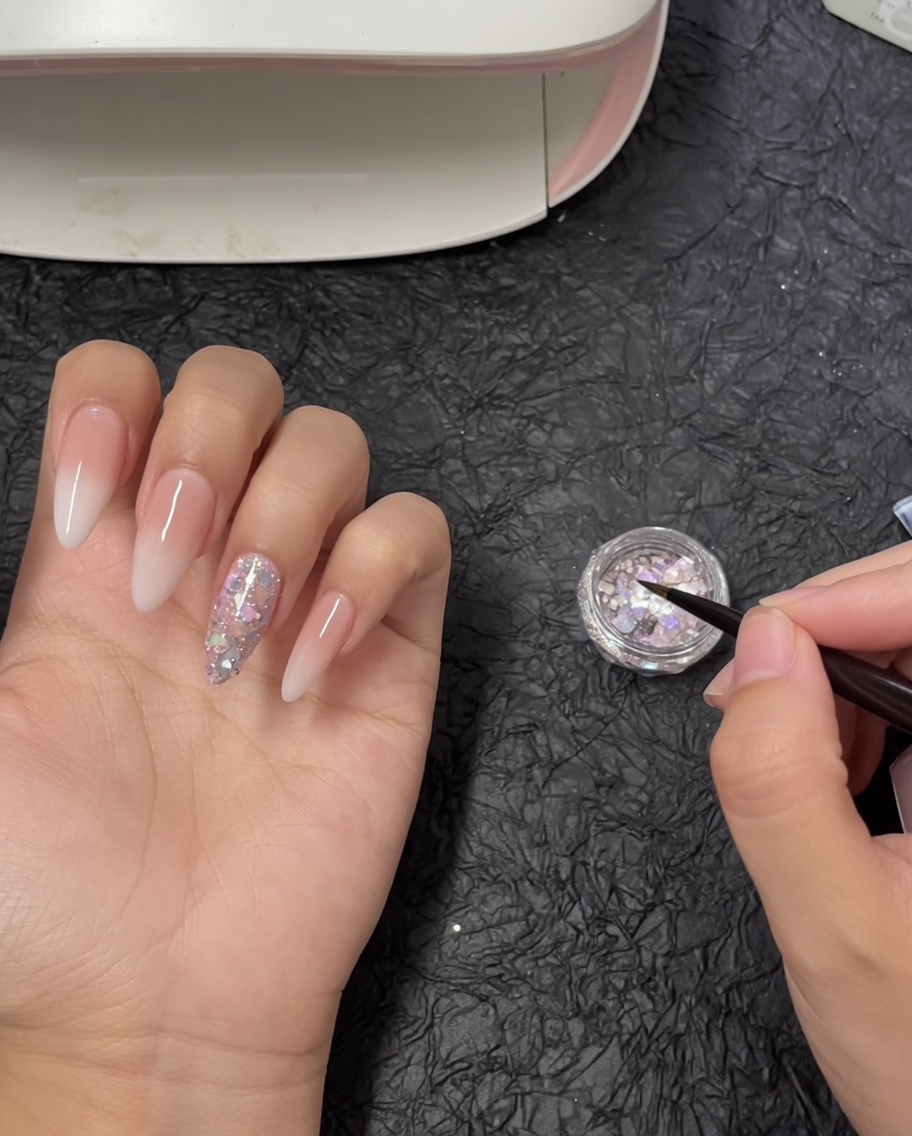 Un pincel de detalle de arte de uñas sumergiéndose en un pequeño tarro lleno de copos de glitter grueso iridiscente.