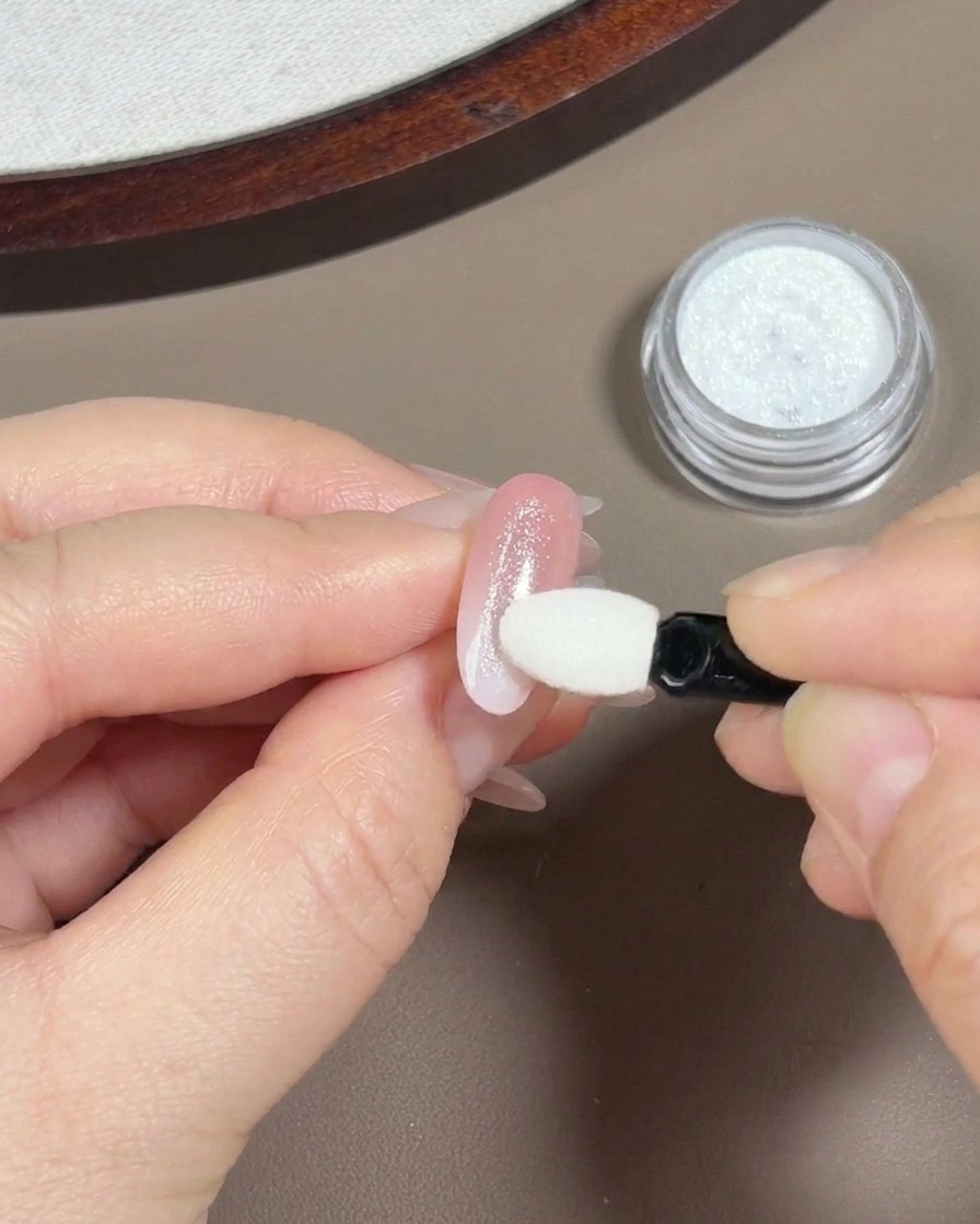 Una mano usando un pequeño aplicador de esponja de sombra de ojos blanco para frotar polvo aurora iridiscente sobre una uña con degradado rosa claro.