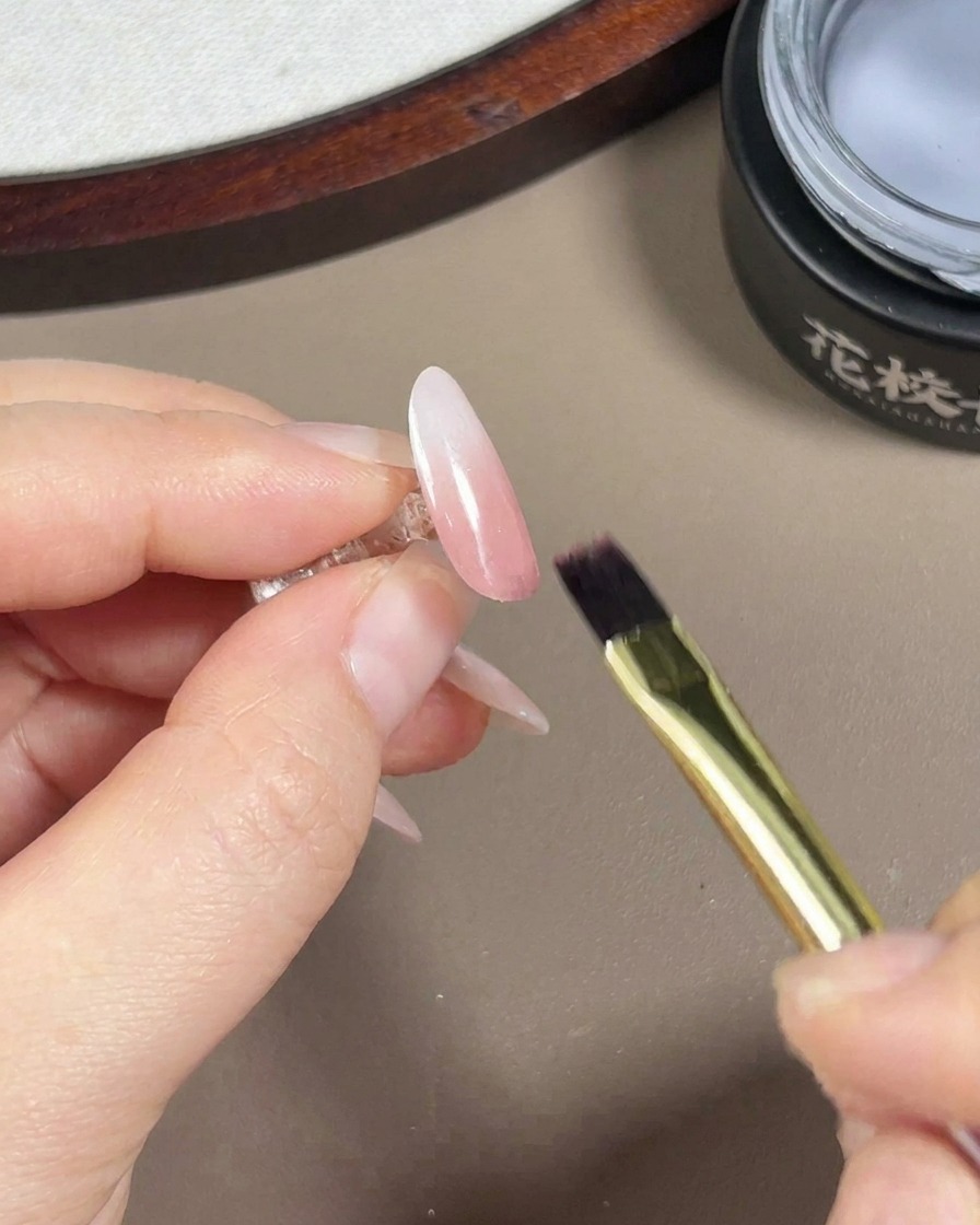 Un pincel para uñas de gel aplicando un top coat transparente y brillante sobre una uña terminada con degradado rosa y blanco con un acabado aurora.