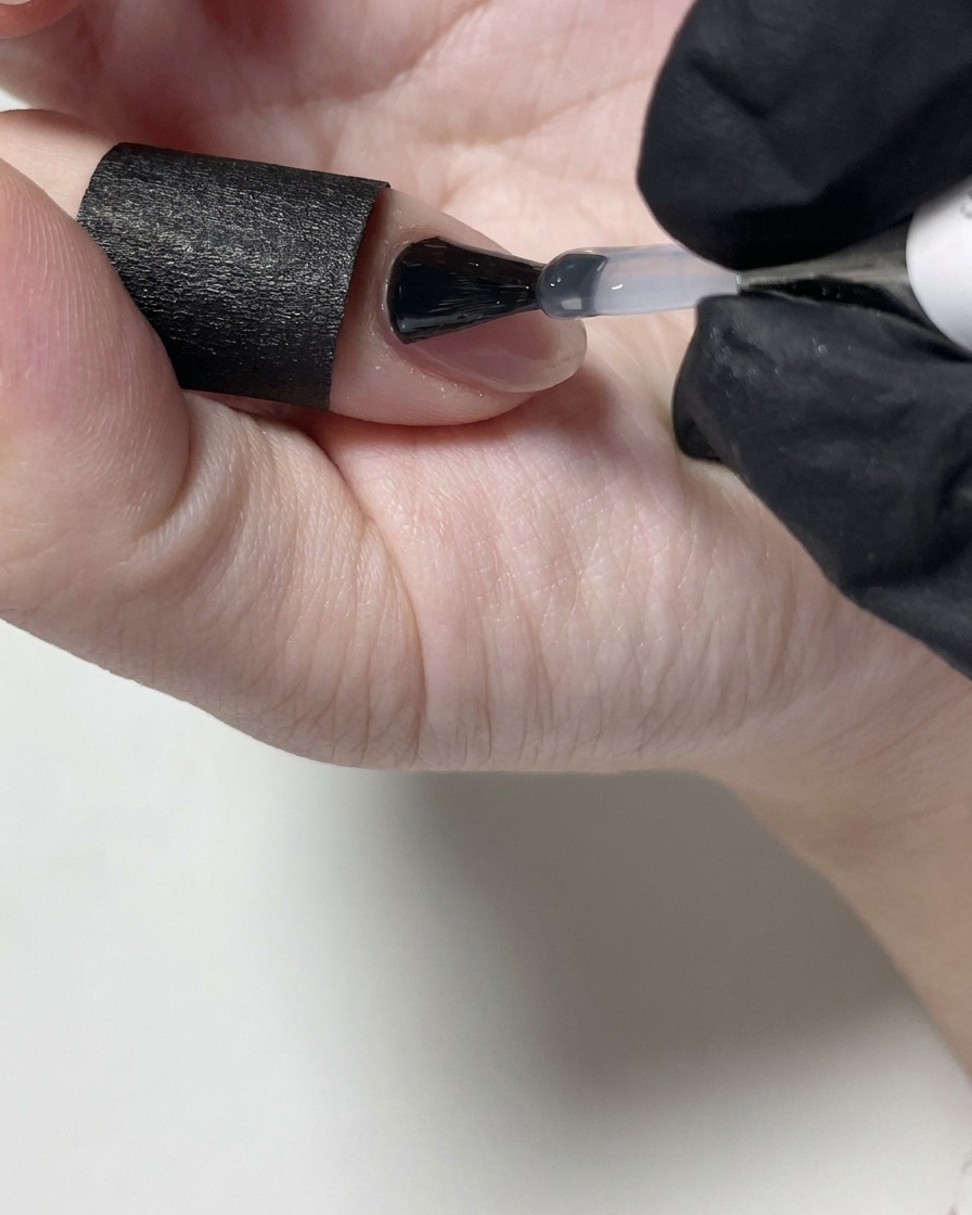 Una técnica de uñas aplicando un top coat mate transparente sobre una uña de gel nude con un pincel negro.