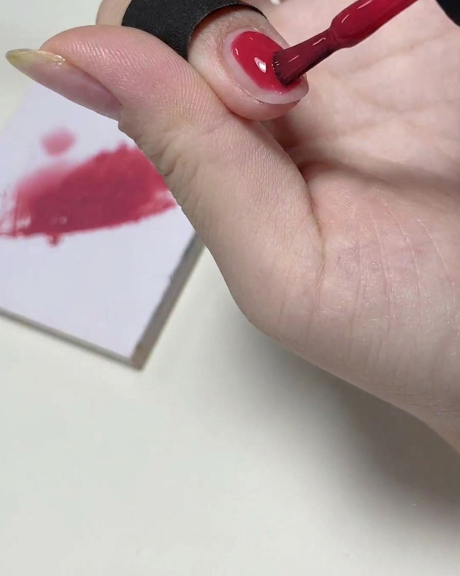 Un pincel de gel aplicando una capa translúcida de esmalte rosa en la mitad superior de una uña, con una paleta de mezcla de fondo.