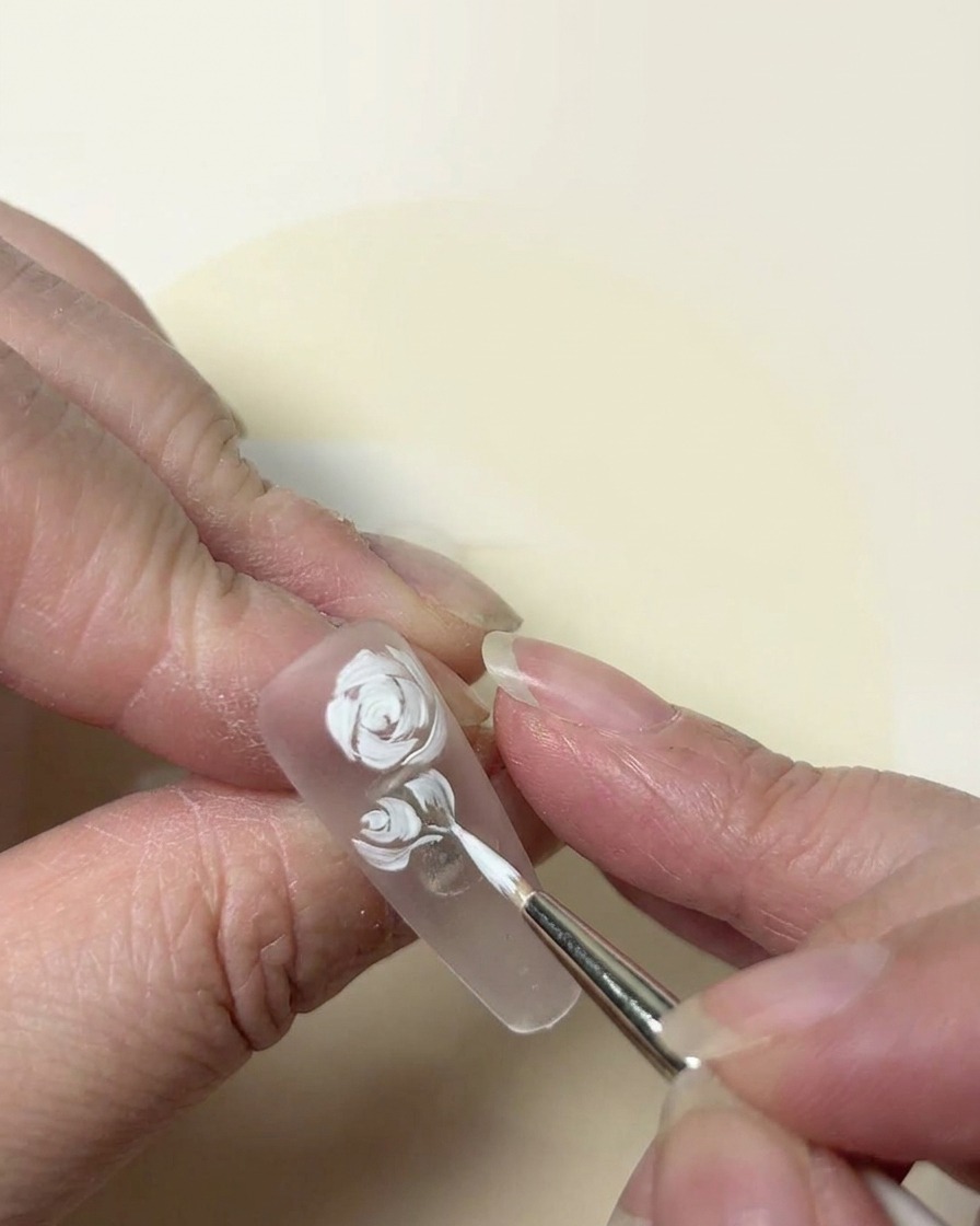 Primer plano pictórico de un diseño de rosa blanca siendo refinado con un pincel de punta fina sobre una uña transparente.