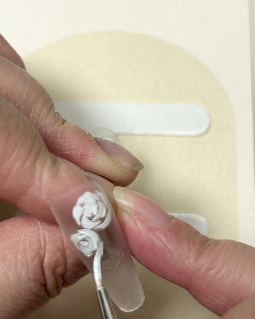 Primer plano de una flor de cinco pétalos siendo pintada en gel blanco al lado de un diseño de rosa establecido.