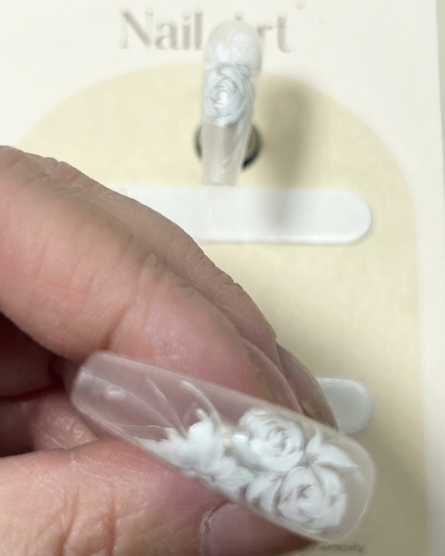Primer plano de una uña con un diseño detallado de rosa blanca, mostrando el pincel añadiendo toques finales de resaltado blanco delicado.
