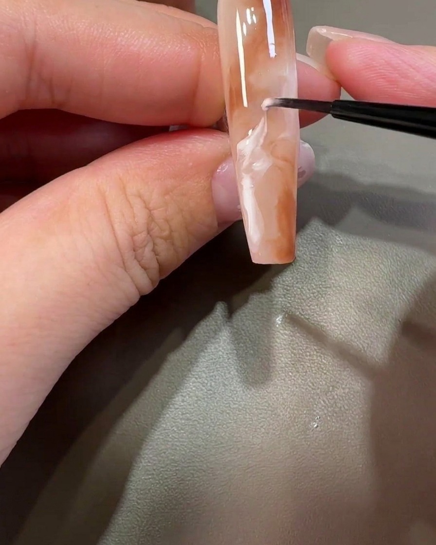 Un pincel liner arrastrando gel blanco a través del diseño marrón para crear patrones de mármol.