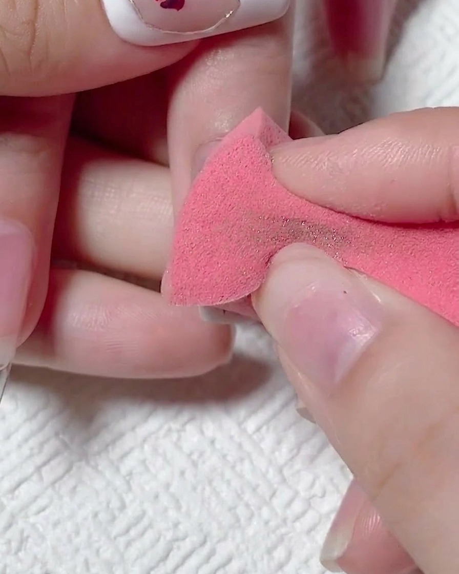 Una esponja de maquillaje rosa retirando el exceso de polvo de cromo de la uña mate.