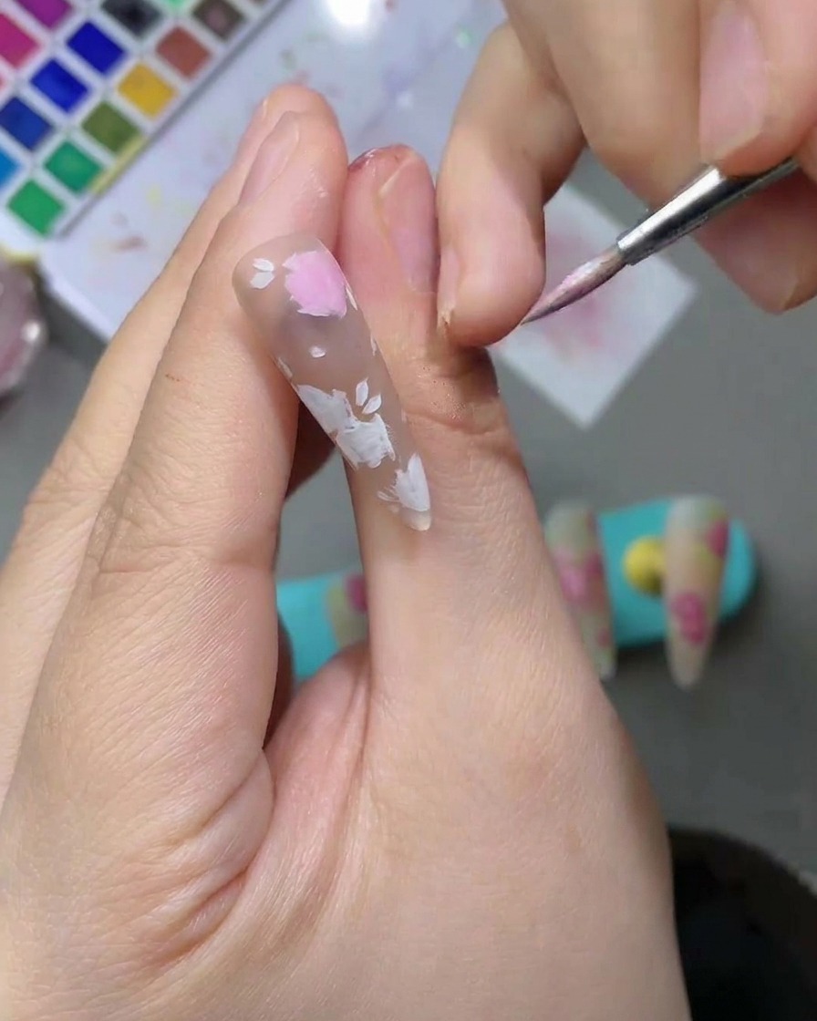 Primer plano de una uña con pétalos de acuarela rosa claro siendo rellenados con un pincel de detalle.