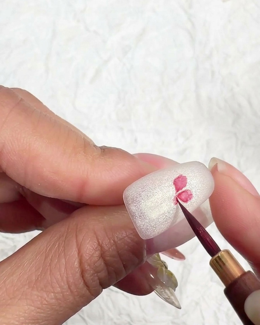 Pincel fino de nail art dibujando el centro de una flor de cerezo rosa sobre una superficie de uña mate.