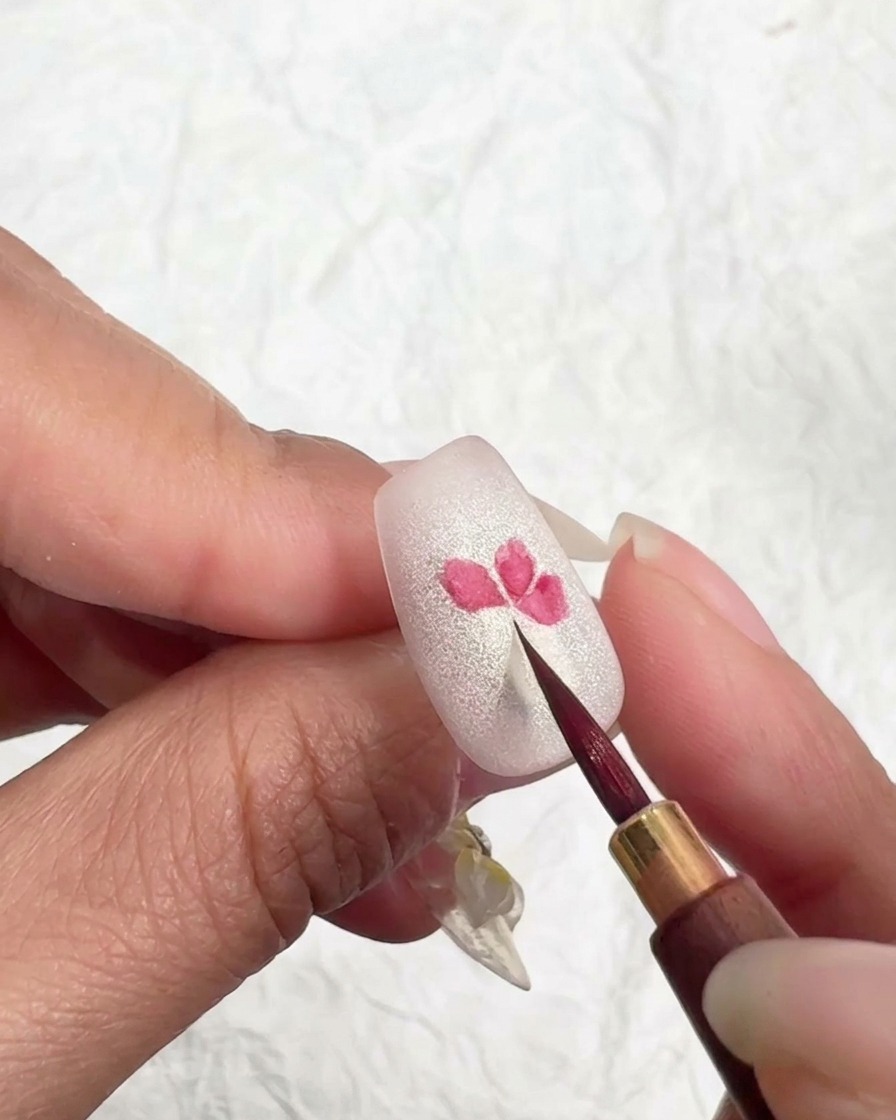 Pincel fino tocando ligeramente los bordes de un pétalo rosa en una uña para suavizar y mezclar el color.