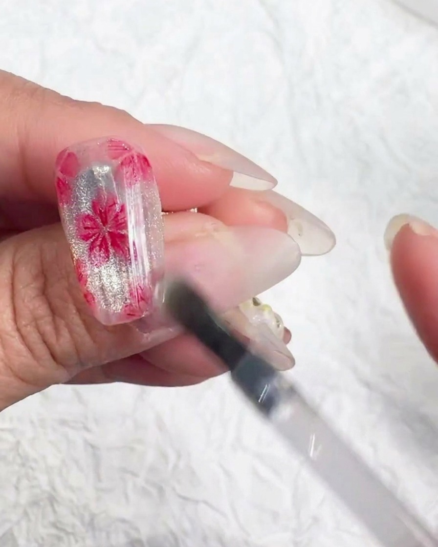 Pequeña herramienta de nail art aplicando diminutos copos iridiscentes en el centro de una flor de cerezo pintada.