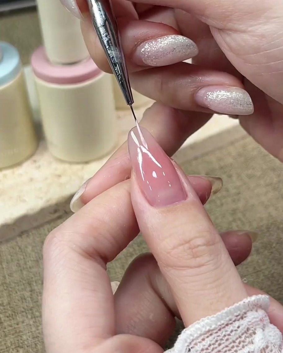 Un pincel de detalle fino dibujando la curva izquierda de la línea de sonrisa francesa con gel rosa pastel.