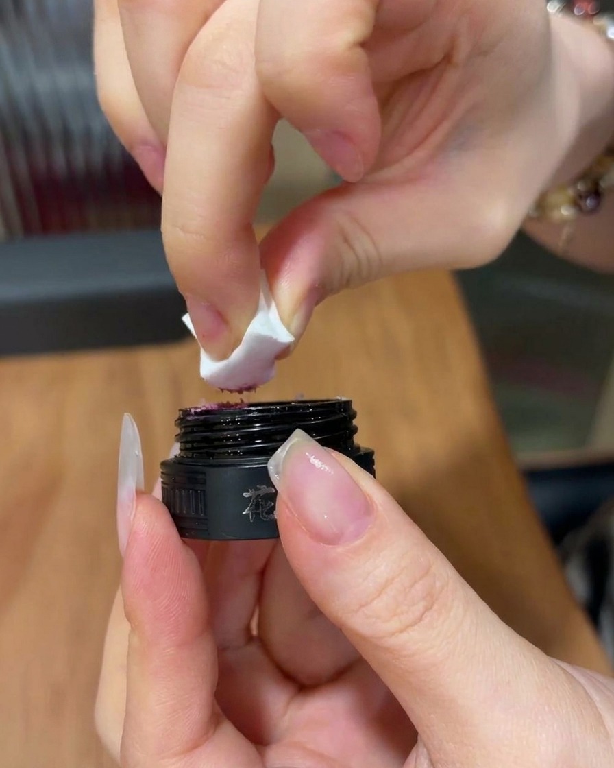 Primer plano de una mano sosteniendo una cuña de esponja blanca, recogiendo una pequeña cantidad de gel rosa brillante de un tarro cosmético negro.
