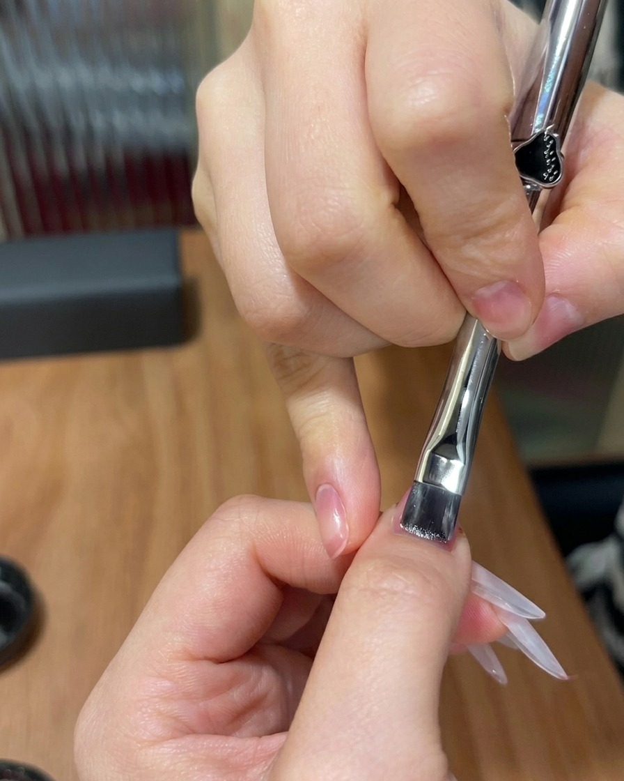 Un pincel plano aplicando una capa fina de gel cat-eye brillante sobre una uña con un degradado rosa preexistente.