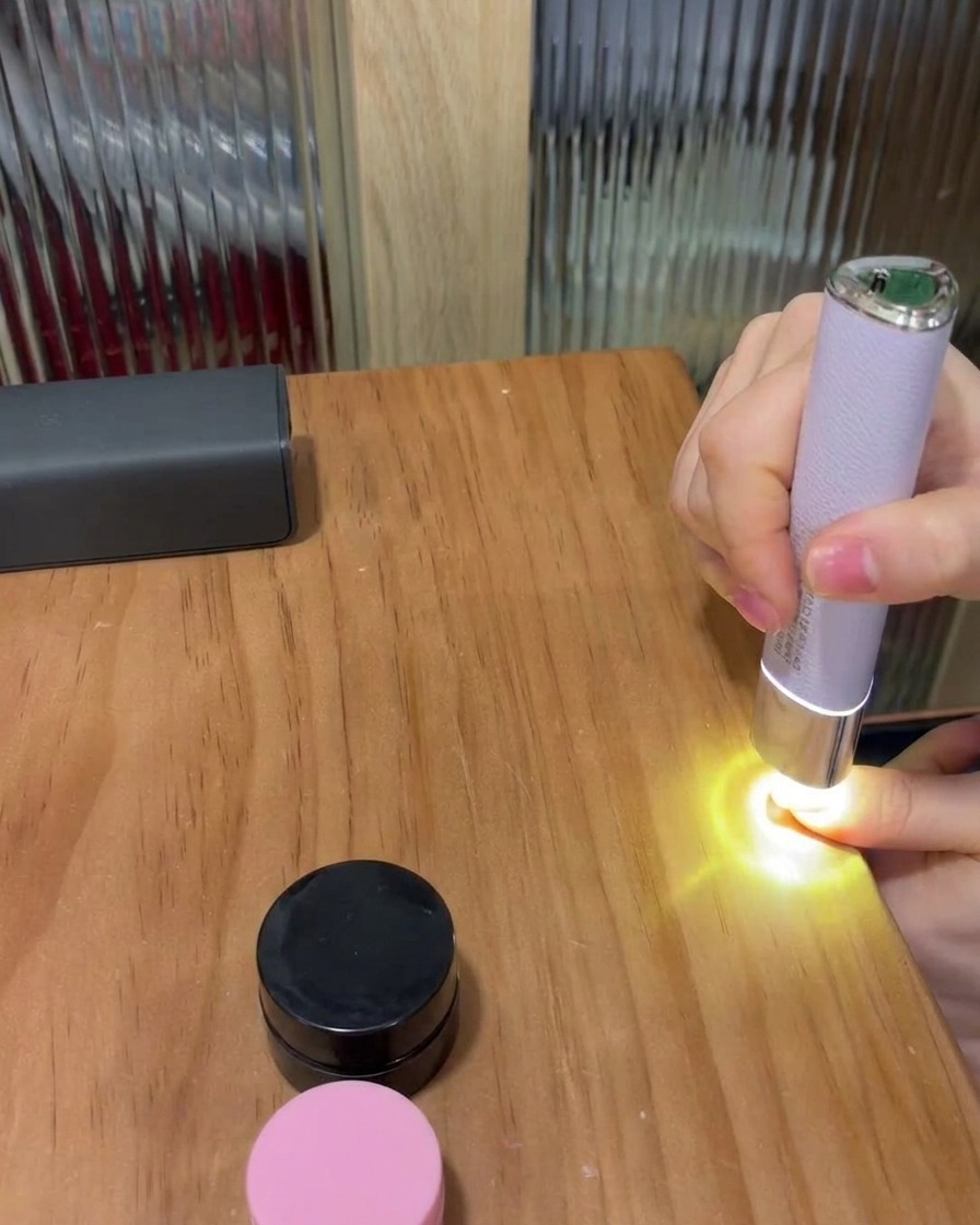 Una uña colocada bajo una pequeña lámpara LED portátil de color morado sobre una mesa de madera, curando el esmalte de gel.