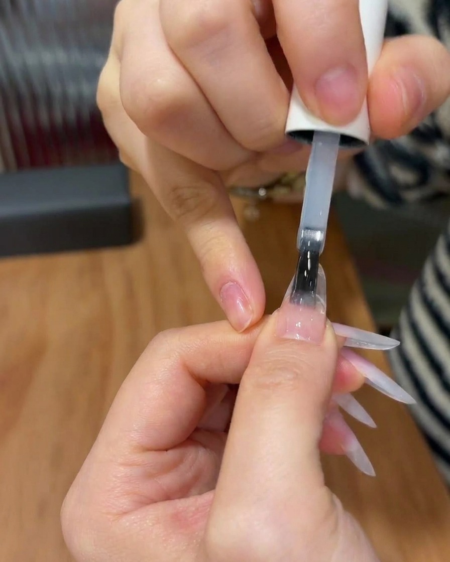 Un pincel de uñas aplicando una capa superior transparente y brillante sobre una uña con un diseño de degradado rosa terminado.