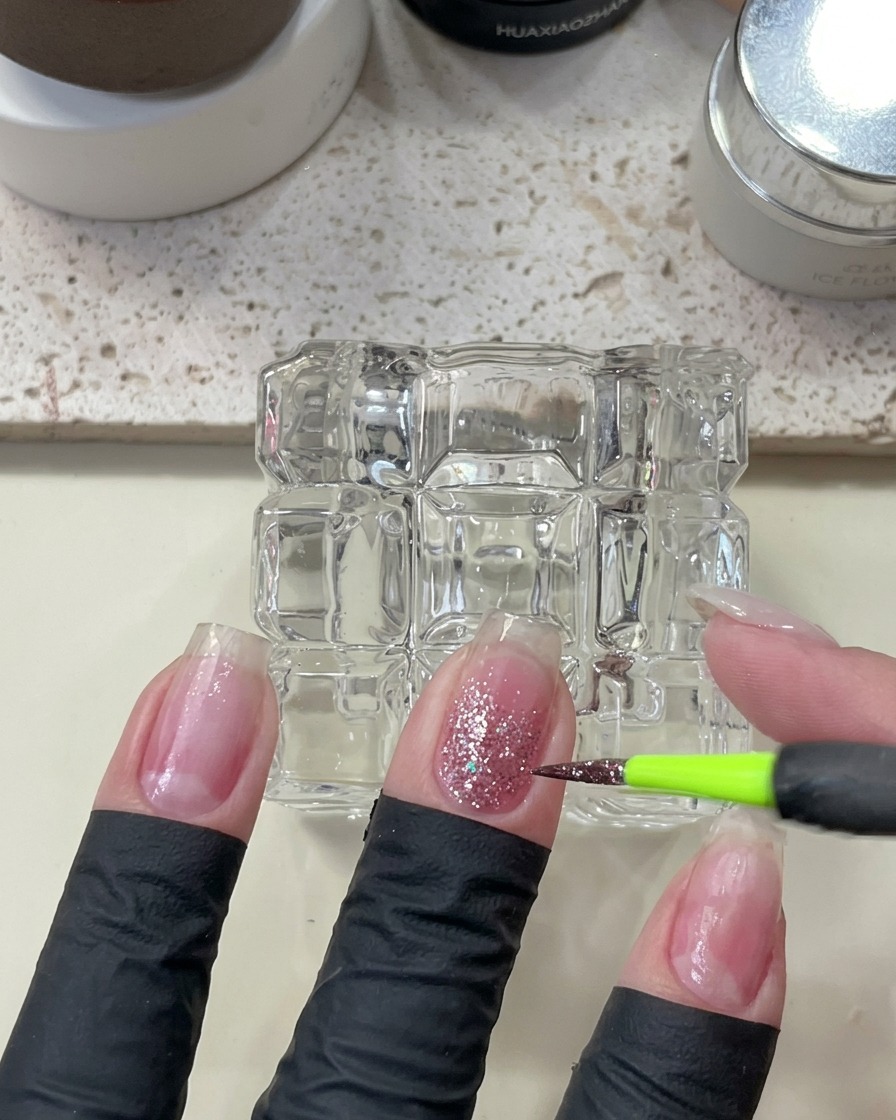 Un pincel fino aplicando glitter rosa brillante sobre la uña para intensificar el efecto.