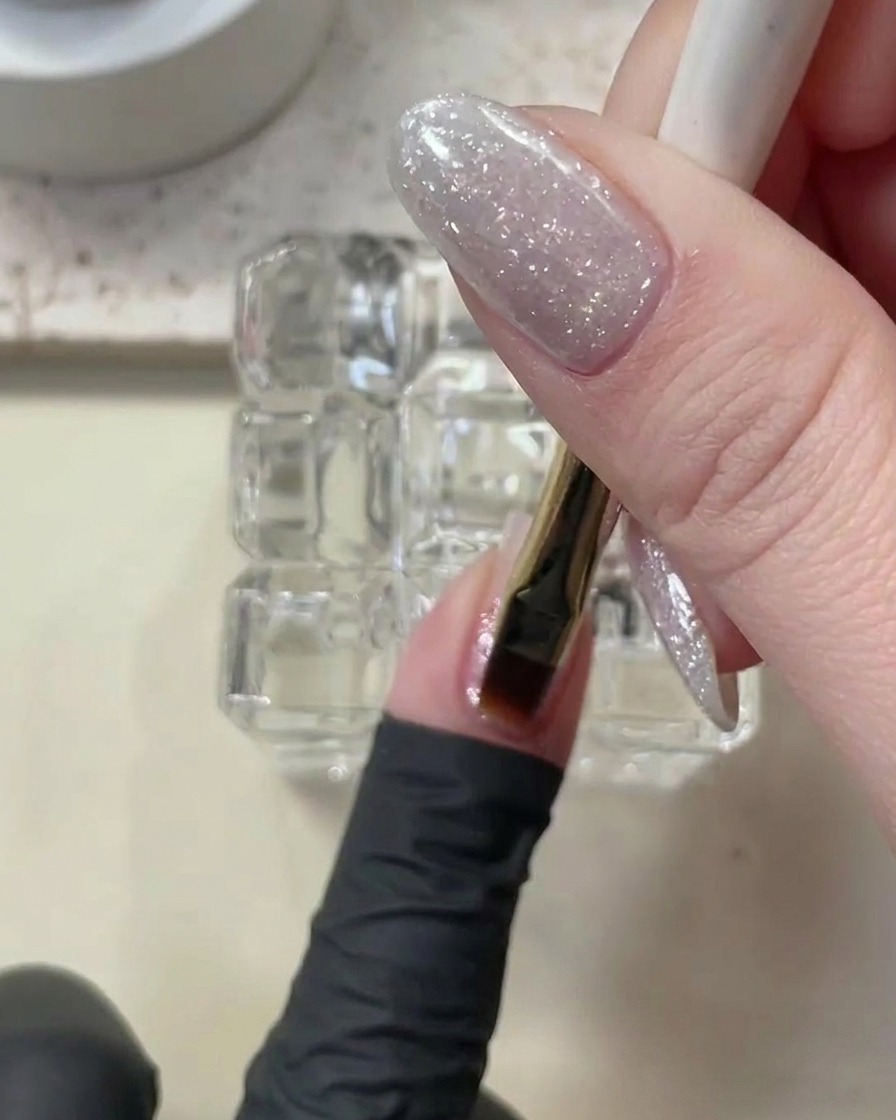 Un pincel aplicando una capa delgada de builder gel transparente sobre la uña con glitter.