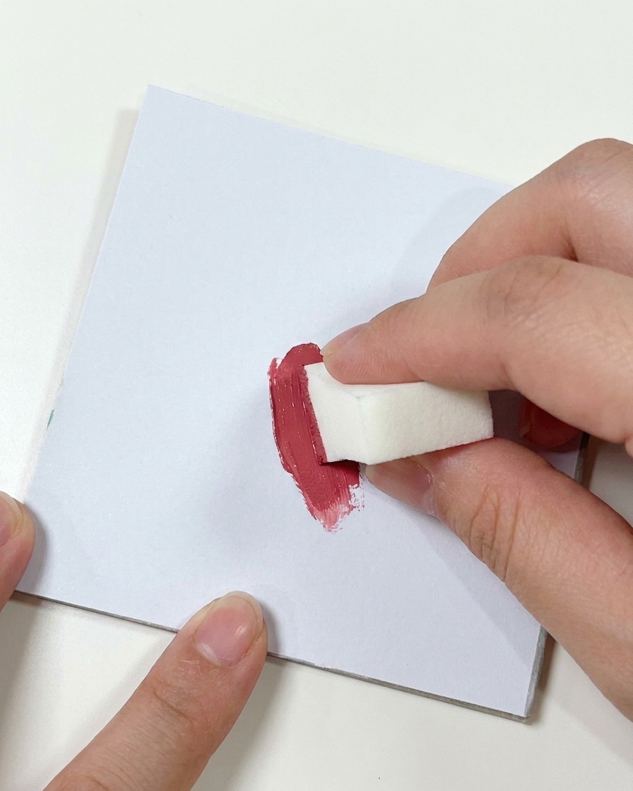 Una mano presionando una pequeña esponja de maquillaje blanca contra una mancha de gel rosa oscuro en una superficie plana.