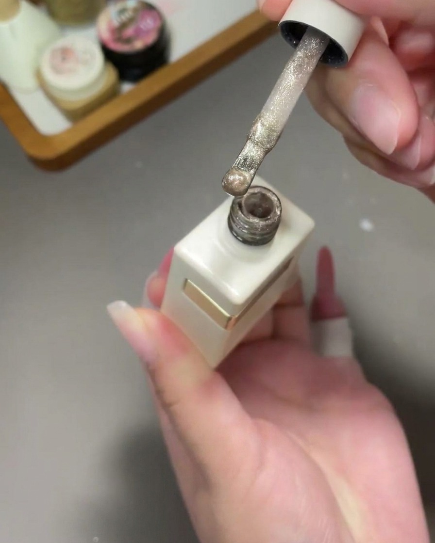 Un pincel aplicando un esmalte de gel ojo de gato dorado brillante sobre una uña con degradado rosa.