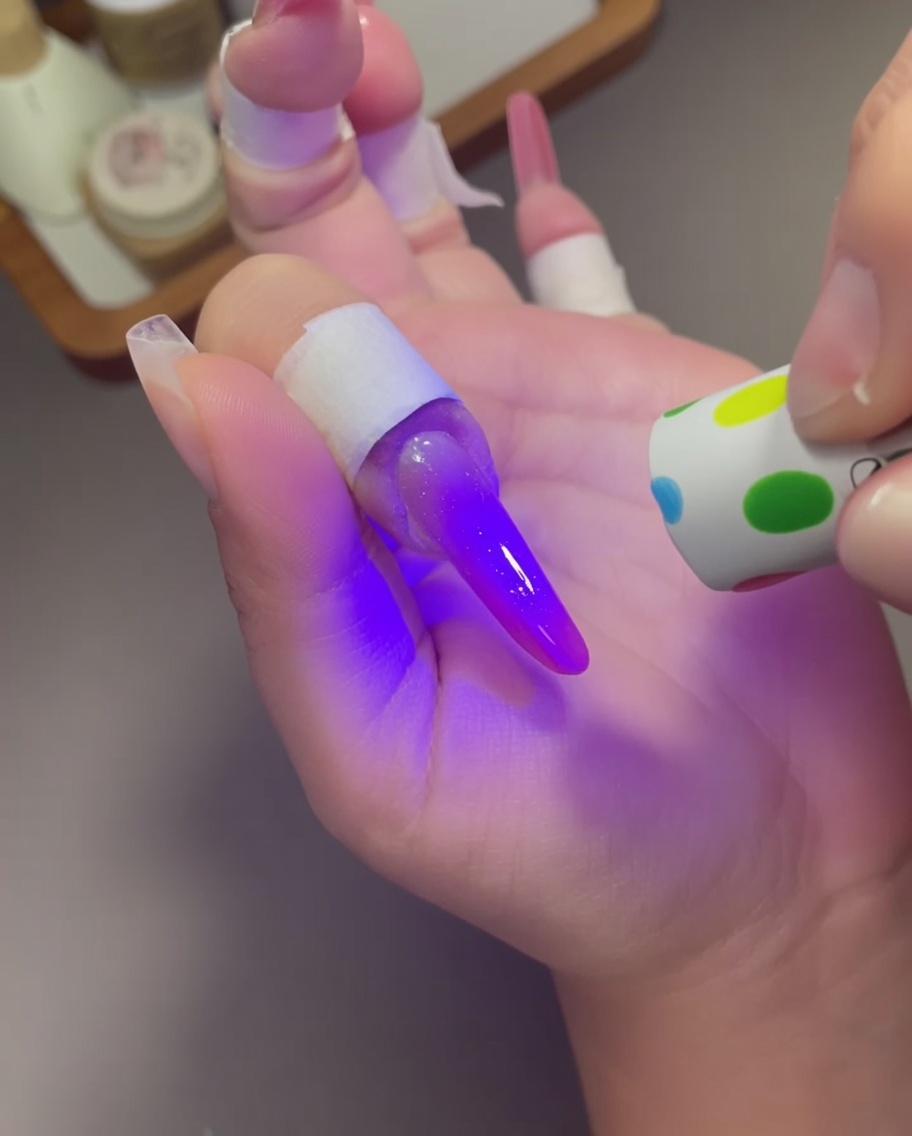Una pequeña lámpara de mano con patrón de puntos sostenida sobre un dedo con un diseño de uñas ojo de gato rosa púrpura brillante.
