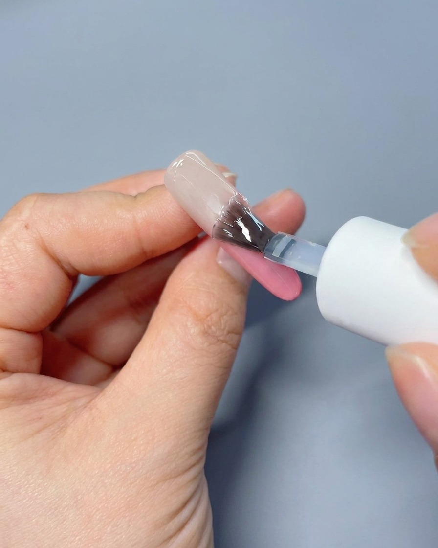 Un pincel aplicando un top coat transparente y brillante sobre una uña con degradado rosa suave.
