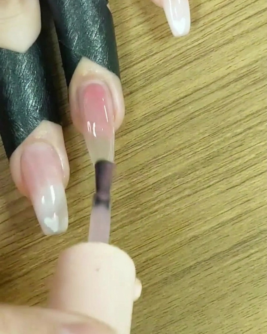 Un pincel aplicando esmalte de gel nude traslúcido en toda la superficie de la uña.