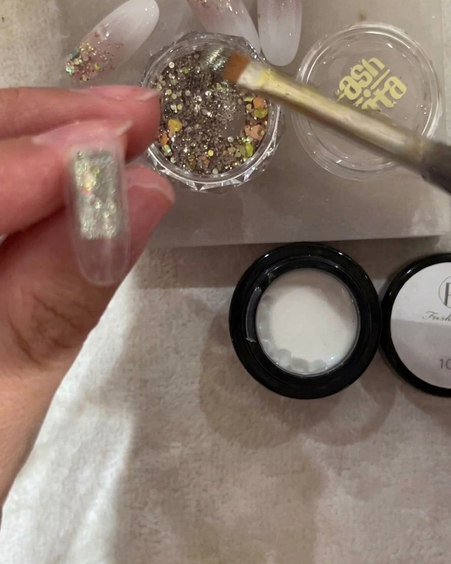 Un pincel aplicando glitter plateado e iridiscente en la mitad superior de un tip de uña transparente.