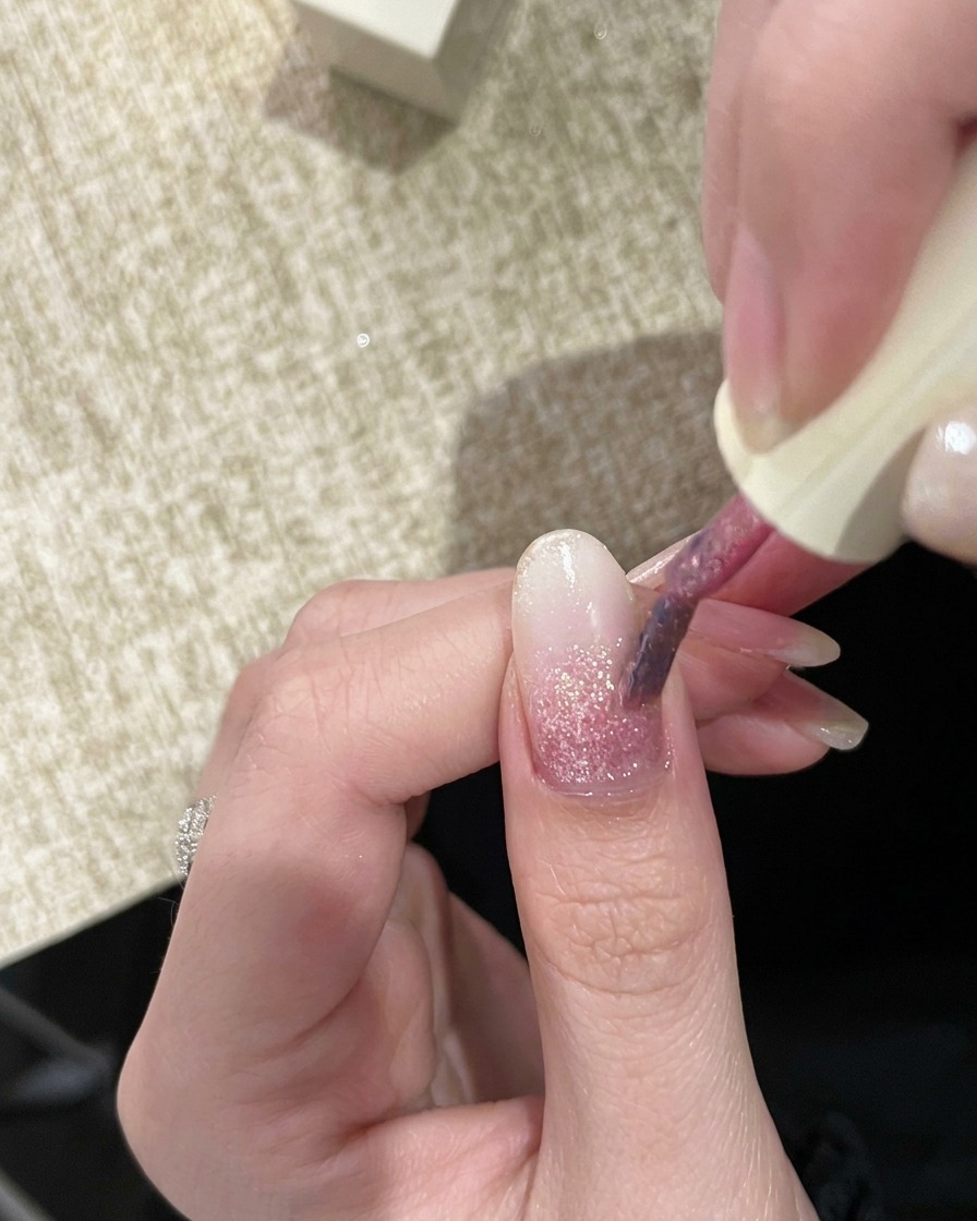Un pincel de uñas aplicando una segunda capa de gel glitter rosa ligeramente más pigmentado.