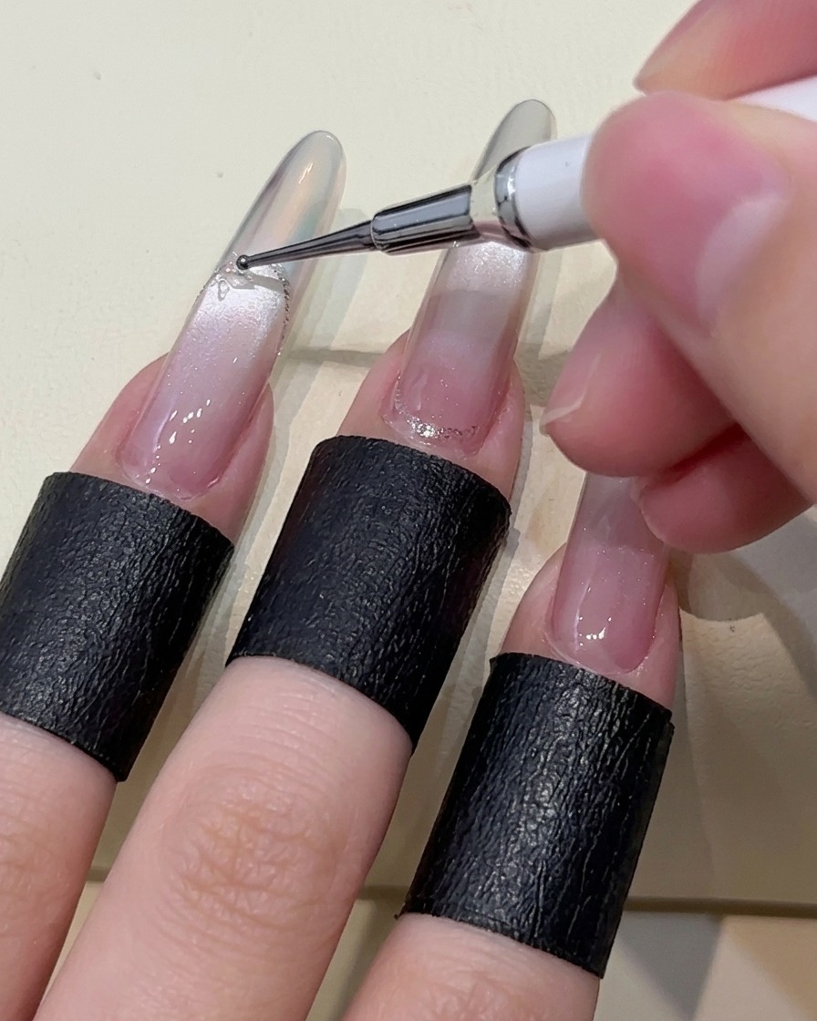 Primer plano de una herramienta de puntos colocando pequeñas gotas de gel adhesivo transparente sobre la superficie de una uña manicurada.
