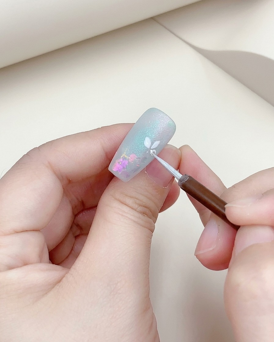 Primer plano de un pincel fino añadiendo un contorno plateado a una flor blanca pintada en una uña.
