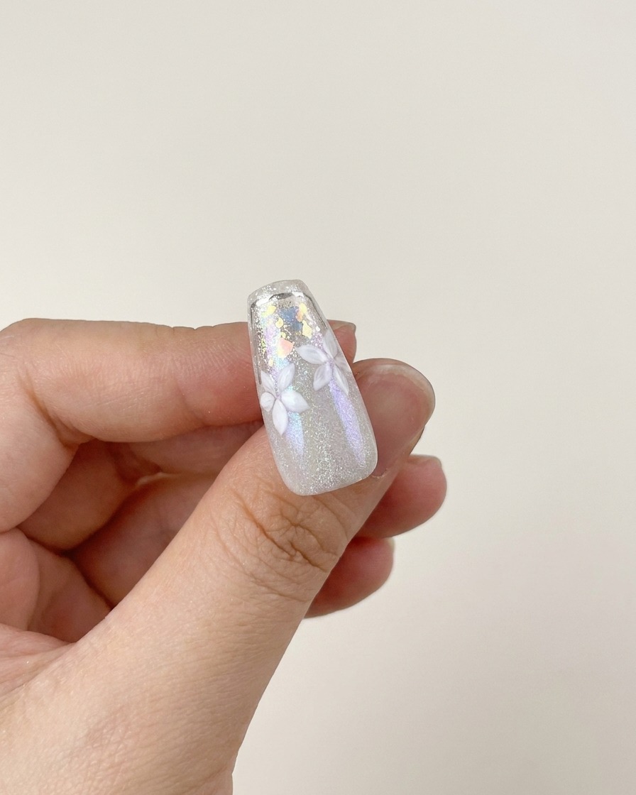Un pincel aplicando una capa de top coat transparente de alto brillo sobre un diseño de uña terminado con flores blancas y escamas plateadas.