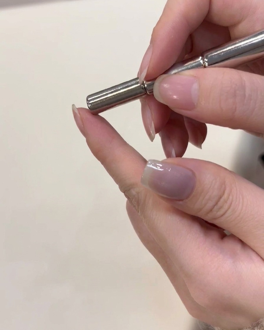 Una herramienta magnética siendo sostenida justo encima de una uña con gel ojo de gato plateado recién aplicado para dar forma a las partículas.