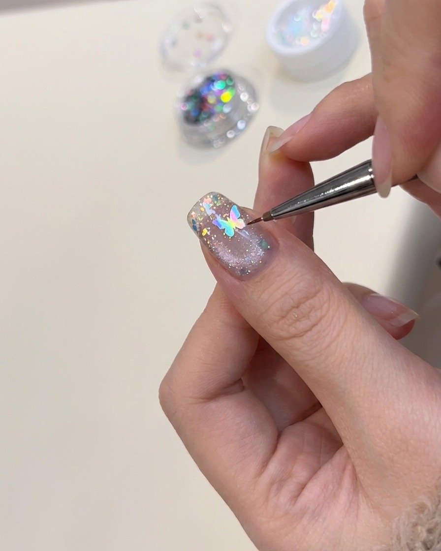 Un par de pinzas posicionando un pequeño dije de mariposa holográfico sobre una uña decorada.
