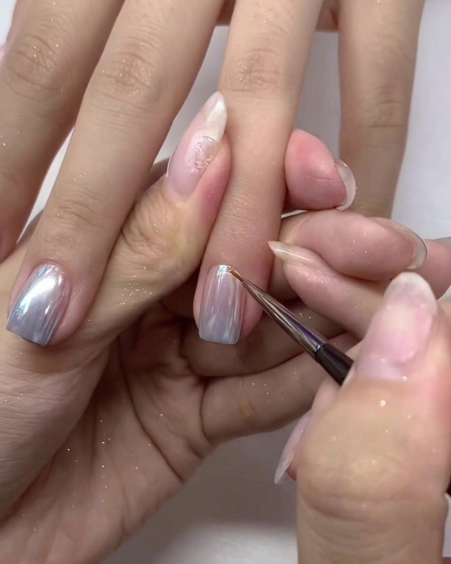 Pincel pequeño aplicando cuidadosamente una última capa de top coat sobre las uñas cromadas plateadas.