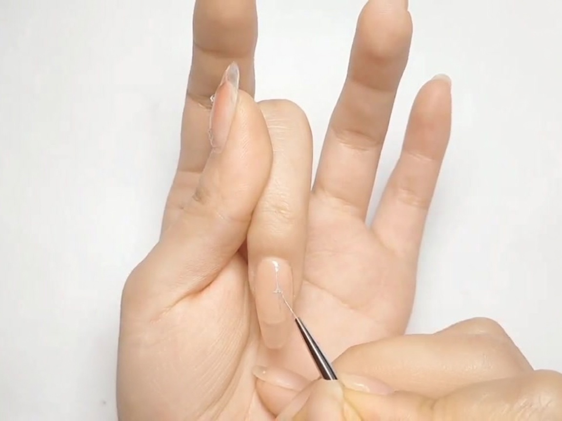 Un pincel fino de nail art dibujando una forma de cruz plateada sobre una uña pintada de nude.