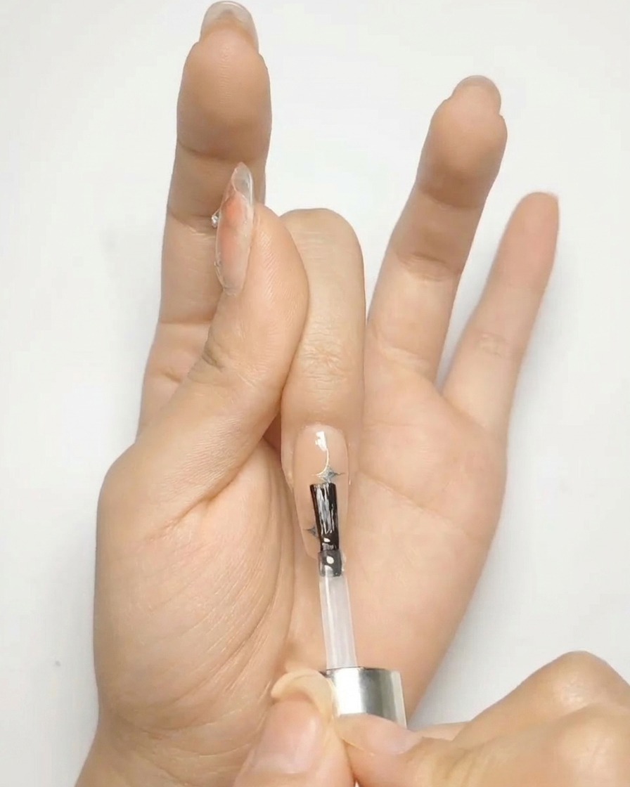 Un pincel aplicando un top coat transparente y brillante sobre una uña de gel nude decorada con estrellas plateadas.