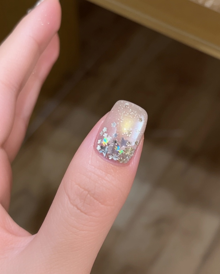 Primer plano de un dedo con lentejuelas grandes en forma de estrella plateada siendo colocadas sobre una uña con gel con un pincel de punta fina.