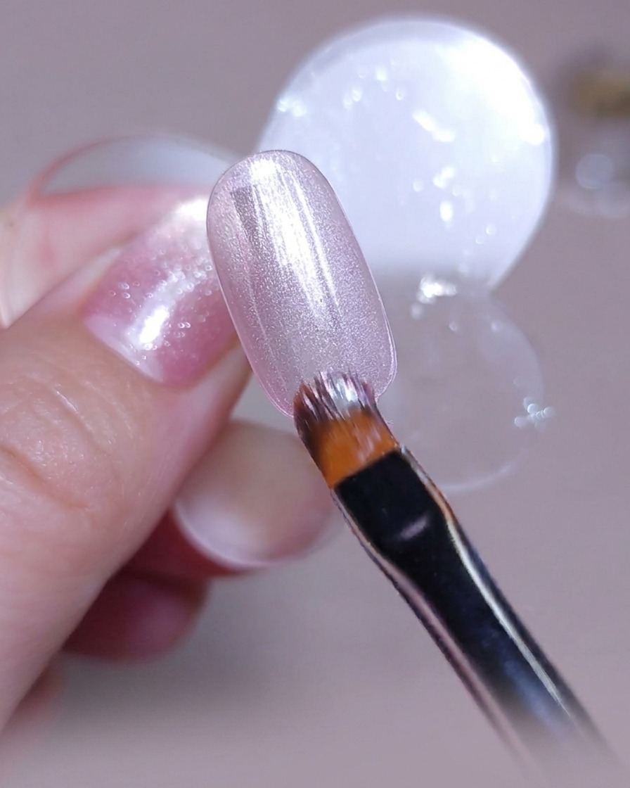Un pincel aplicando una capa uniforme de esmalte en gel ojo de gato rosa plateado sobre un tip de uña transparente.