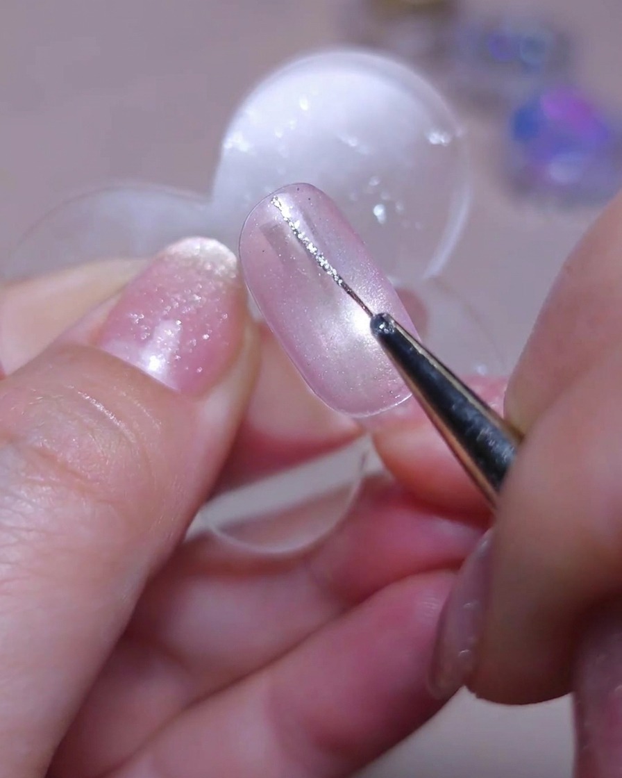 Un pincel fino de detalle dibujando una delicada línea vertical con gel de glitter plateado en el lado izquierdo de una uña ojo de gato.
