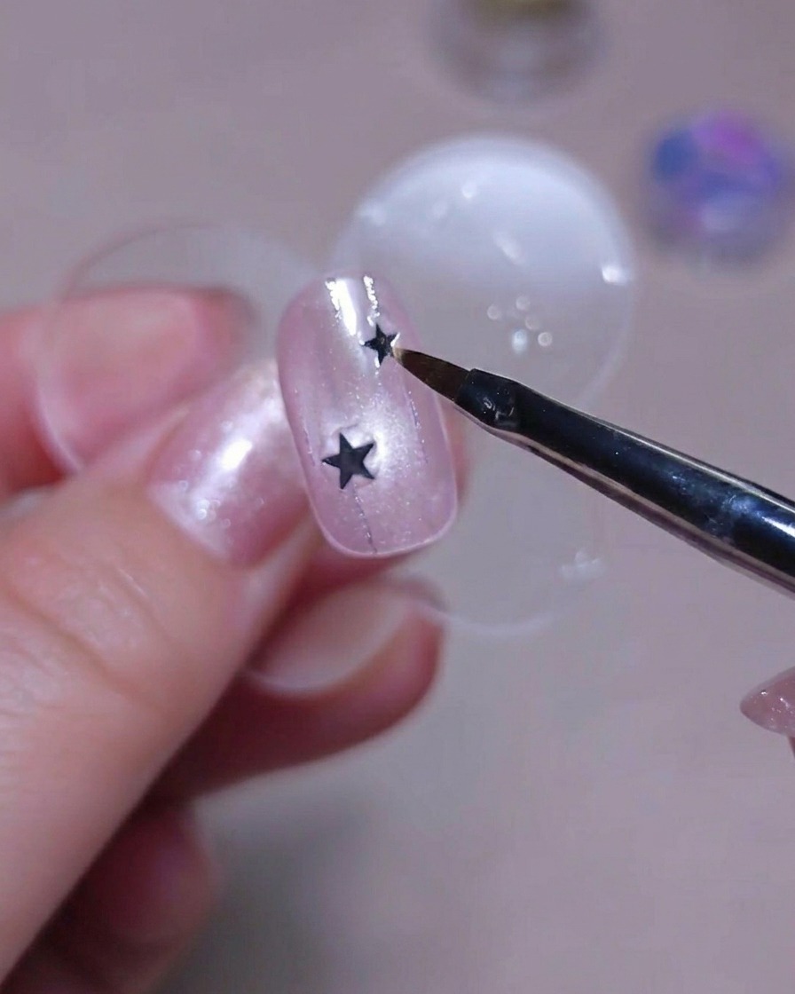 Un primer plano de la punta de un pincel sosteniendo una pequeña lentejuela de estrella plateada sobre una uña rosa con glitter.