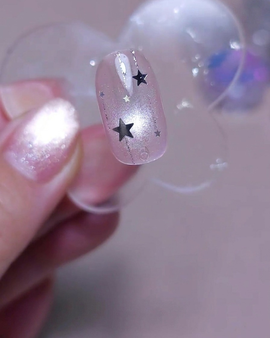 Un pincel aplicando una capa final de top coat transparente y brillante sobre una uña decorada con líneas verticales y lentejuelas de estrellas plateadas.