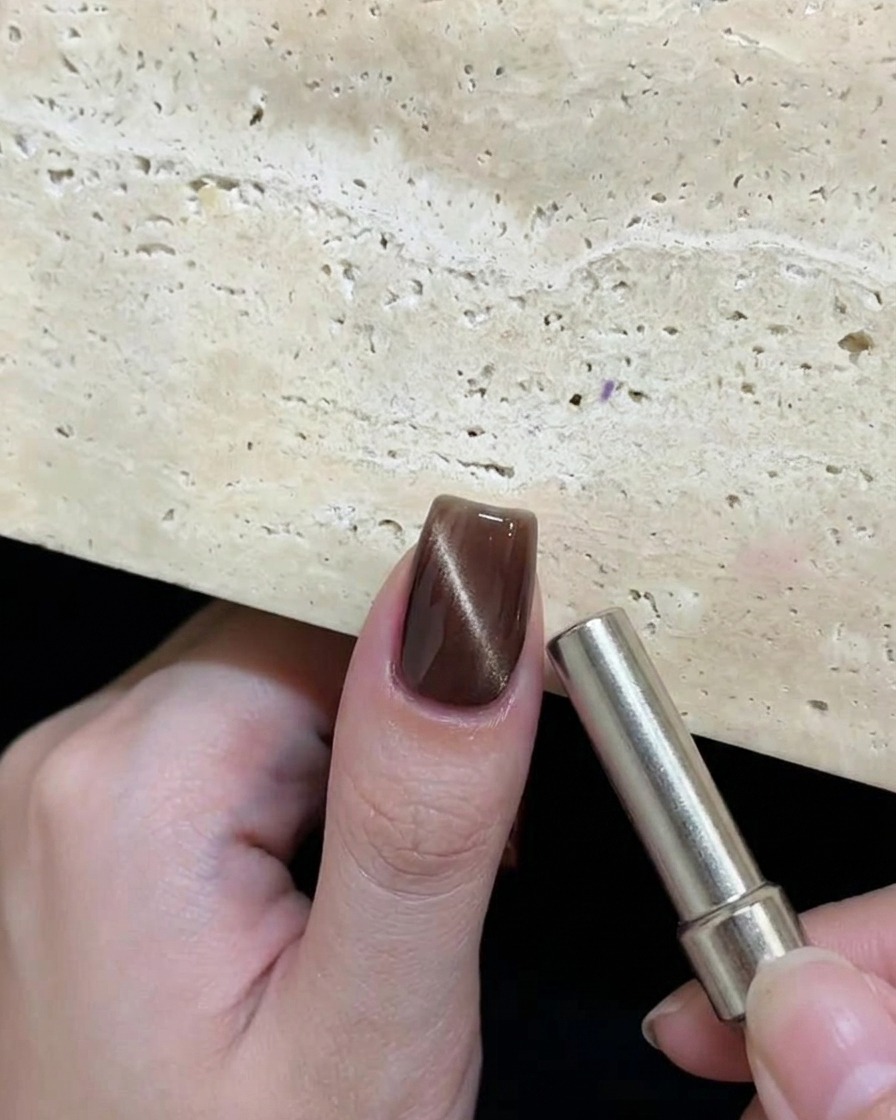 Una mano sosteniendo un imán cilíndrico cerca de una uña con gel cat eye marrón húmedo para crear un patrón reflectante circular.