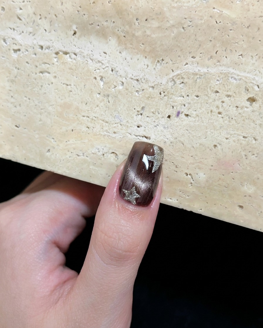 Una uña con gel cat eye marrón decorada con dos estrellas de plata platino pintadas a mano.