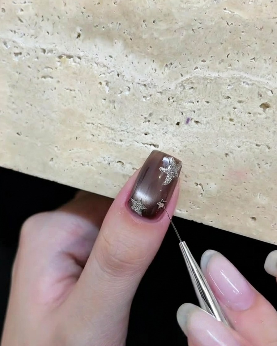 Una herramienta colocando piezas de glitter de estrellas plateadas sobre una uña de gel marrón.