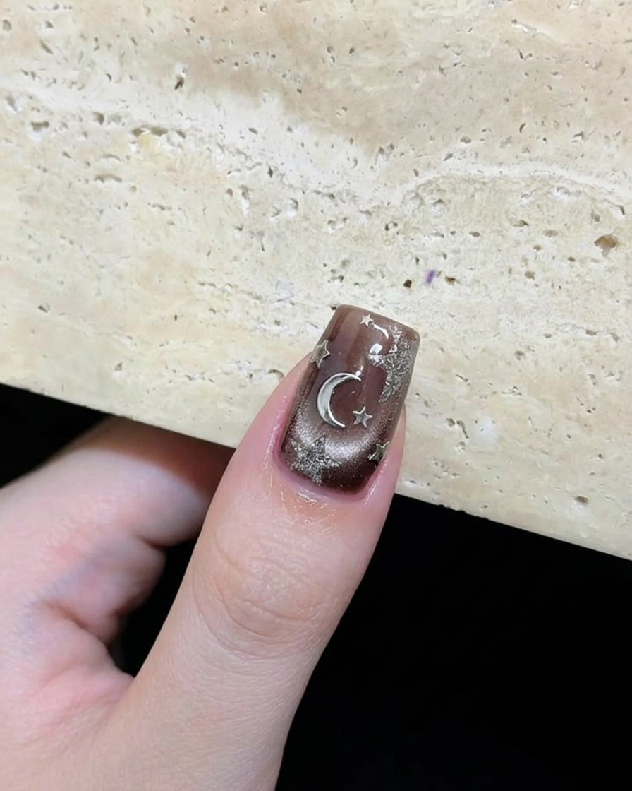 Primer plano de una uña marrón oscuro terminada con estrellas plateadas y un acento de luna dispuestos artísticamente.