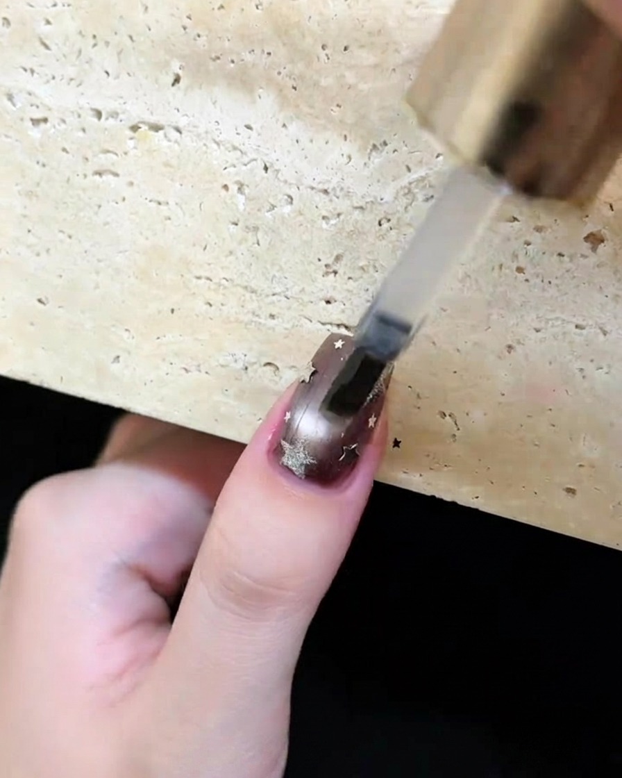 Un pincel aplicando un top coat transparente y brillante sobre una uña marrón oscuro decorada con estrellas y lunas plateadas.