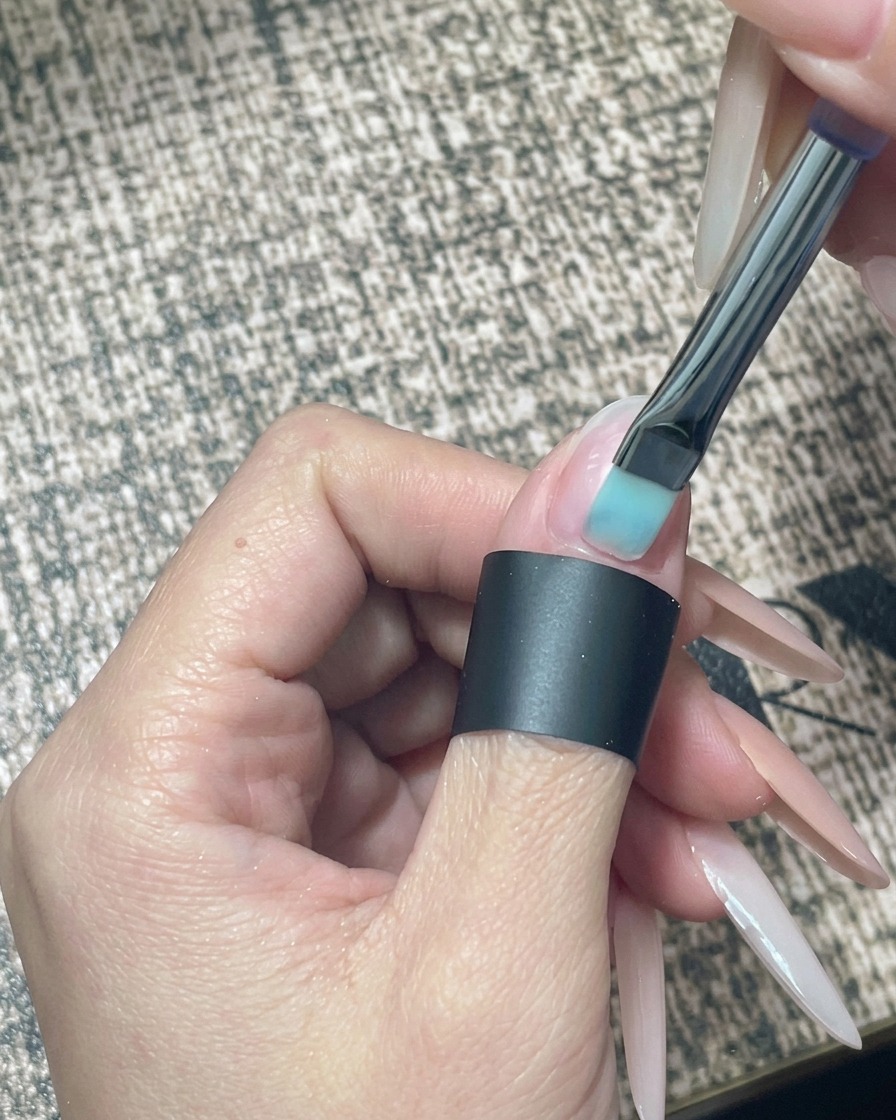 Primer plano de un pincel aplicando esmalte de gel nude translúcido sobre una uña natural preparada.